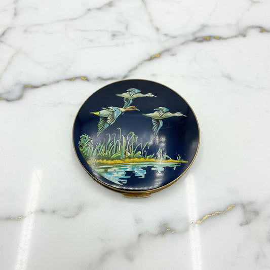 beautiful enamel powder compactfeaturing three ducks flying over water on a mirror compact lid on a marble surface