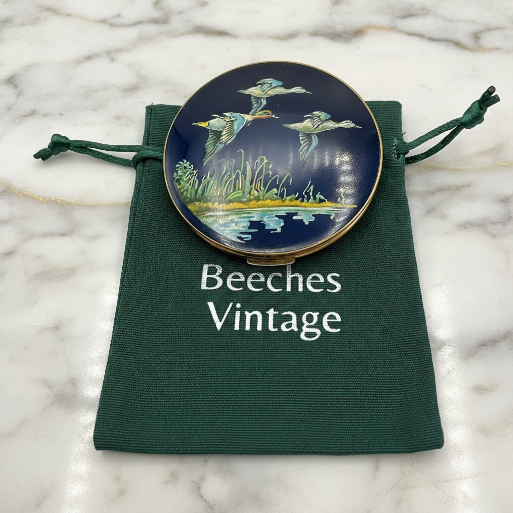 beautiful enamel featuring three ducks flying over water on a mirror compact on a green cotton bag on a marble worktop