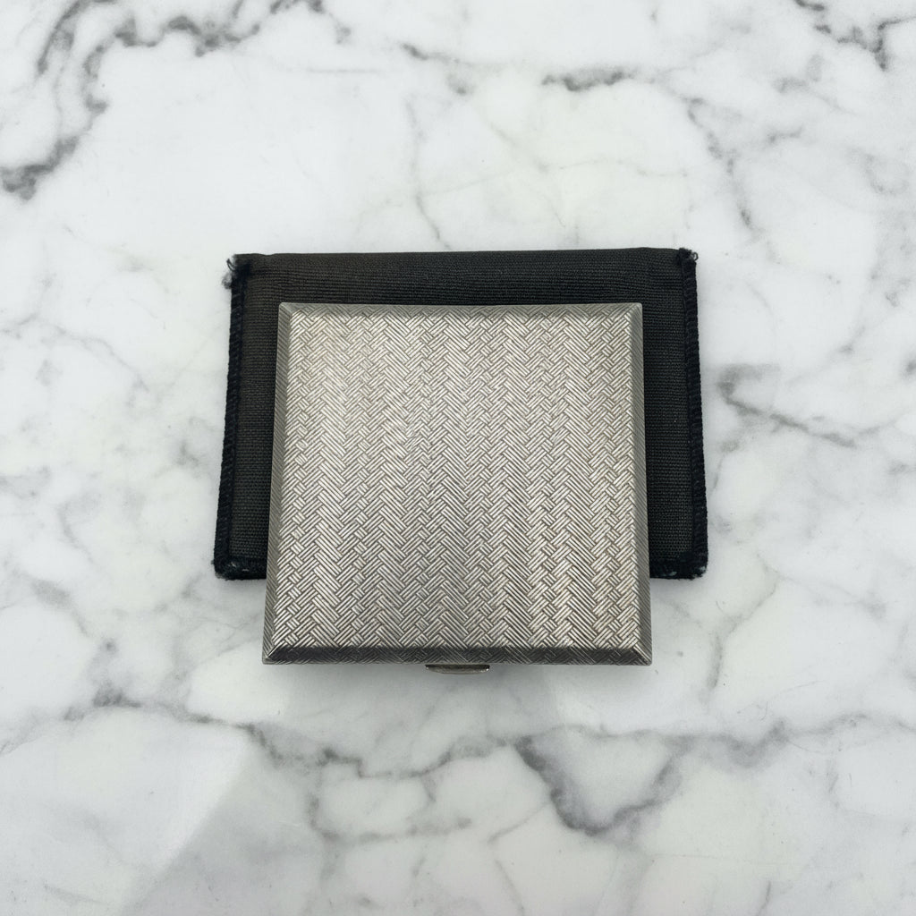 A silver coloured square compact with a woven pattern to the lid. It is sitting on a black bag.