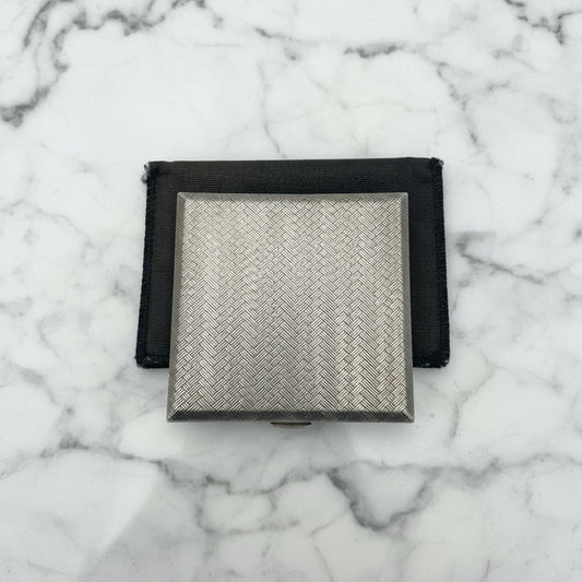 A silver coloured square compact with a woven pattern to the lid. It is sitting on a black bag.