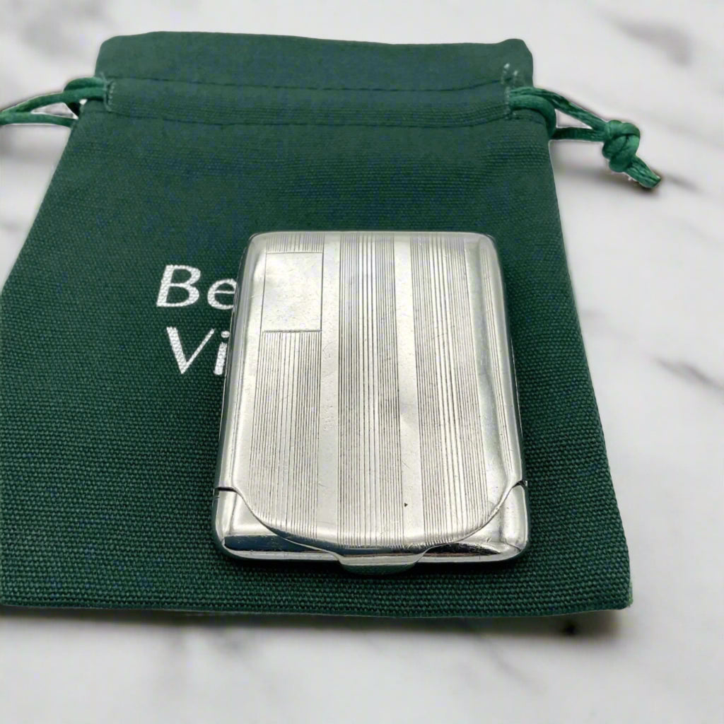 Silver matchbook case with a lined pattern and blank box for engraving initials in left hand corner. Case is on green Beeches Vintage cotton bag.
