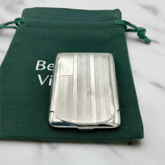 Silver matchbook case with a lined pattern and blank box for engraving initials in left hand corner. Case is on green Beeches Vintage cotton bag.