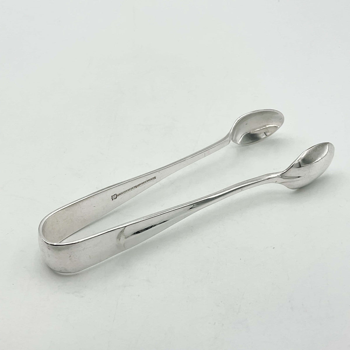 1930s style silver plated sugar tongs with a lined 'rat tail'  design to the arms. The sugar tongs are sitting on a white background.
