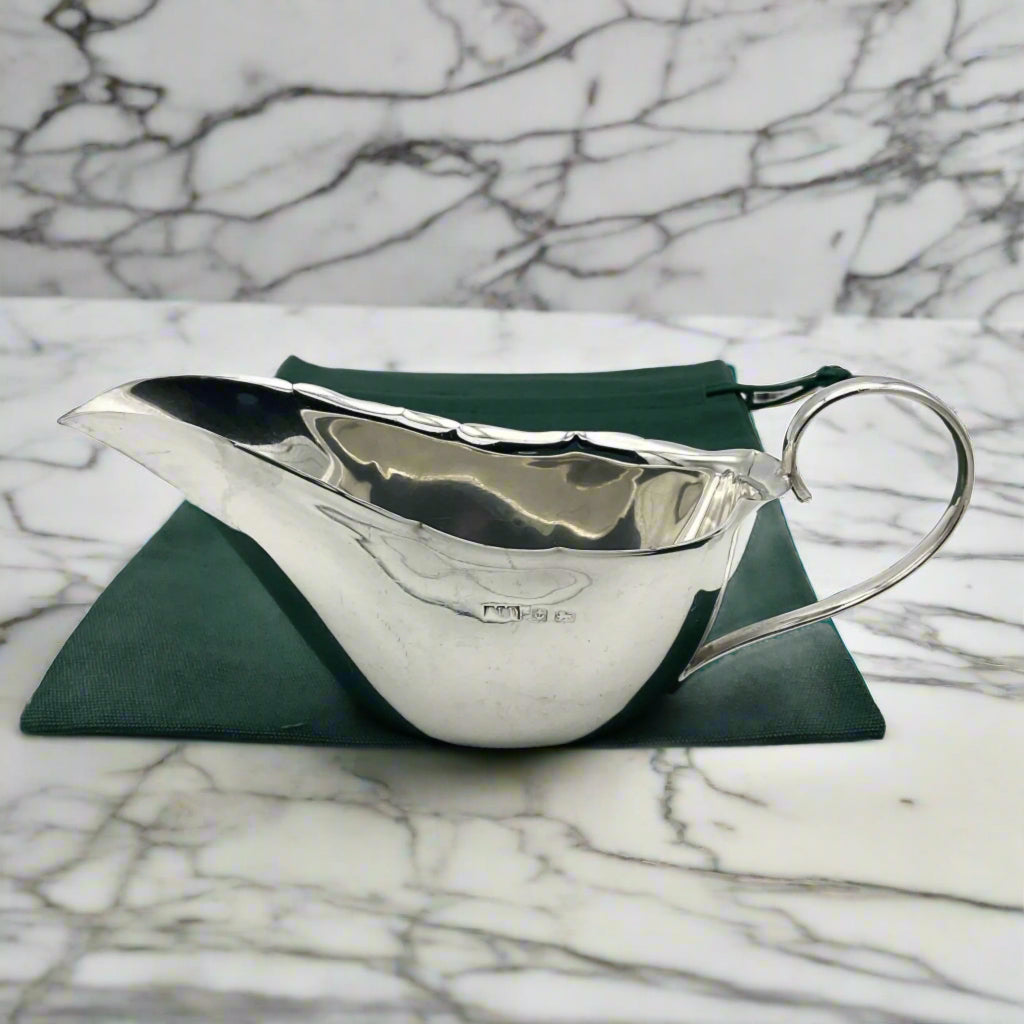 Shiny Silver sauce boat sitting on a green cotton bag. The sauce boat has a loop handle , crimped edges and Silver hallmarks on one side on a marble surface.