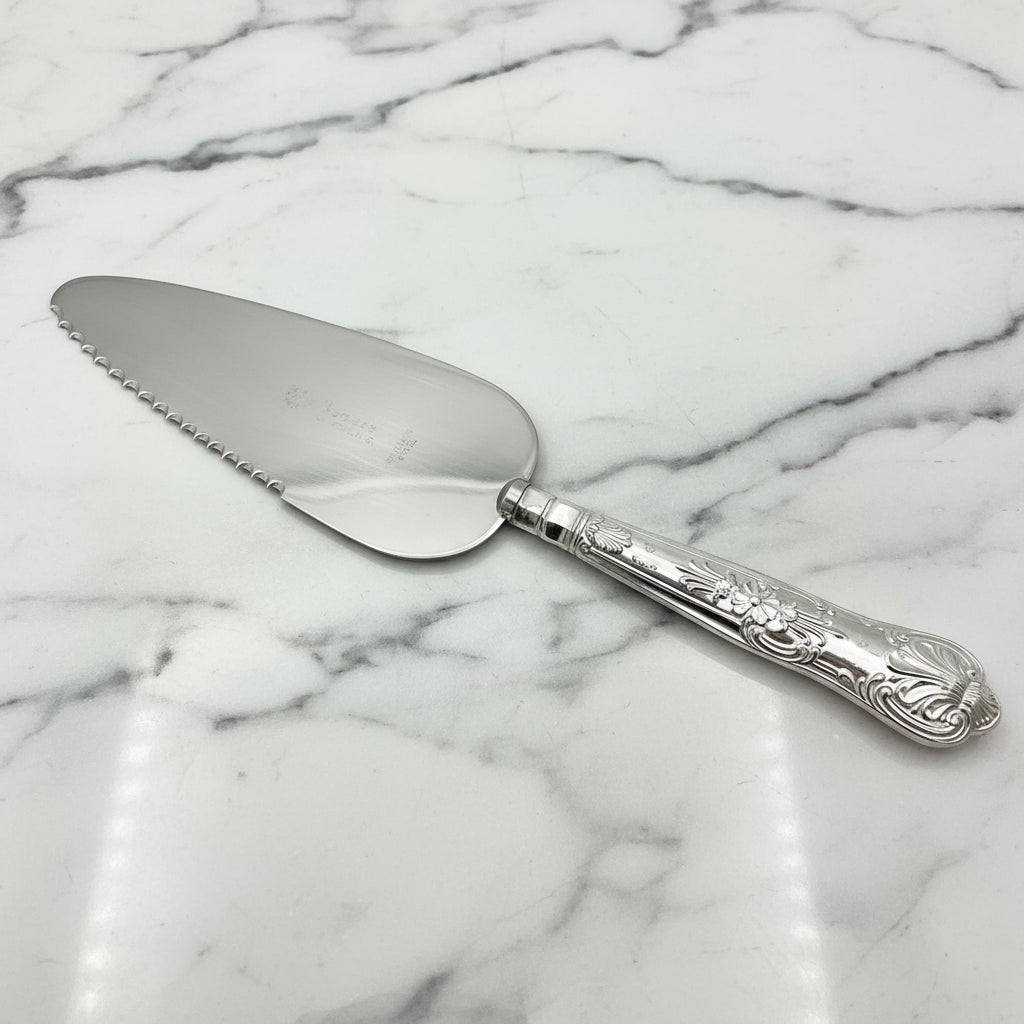 A silver coloured metal cake or pie slice on a marble background. The handle has a Queens pattern elaborate design.