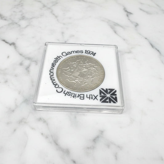 One dollar coin in a white case with 10th British Commonwealth Games 1974 around it.