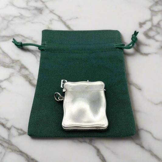 Antique sterling silver Vesta case with a wavy shape on the sides, a ring on one side, and a chain attached. It is placed on a green gift bag with a snap closure.