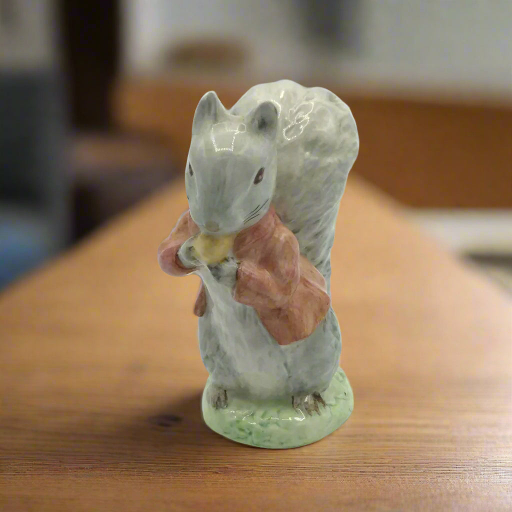 A figurine of a gray squirrel with a big bushy tail wearing a light red jacket, nibbling on a nut. Sitting on a wood shelf