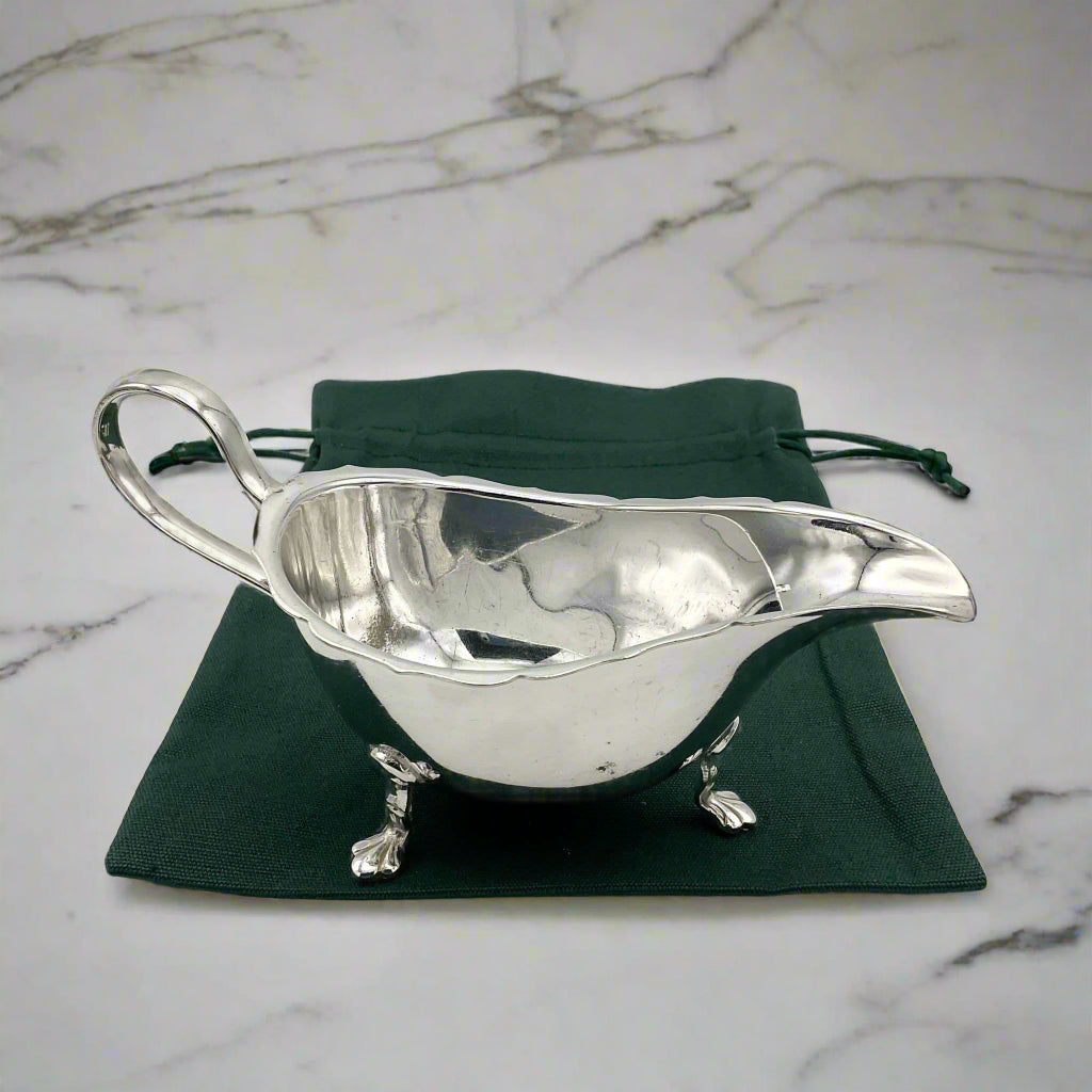 A silver-plated gravy boat with three feet and a loop handle, displayed on a green gift bag on a marble surface