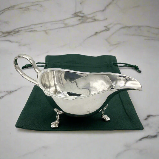 A silver-plated gravy boat with three feet and a loop handle, displayed on a green gift bag on a marble surface