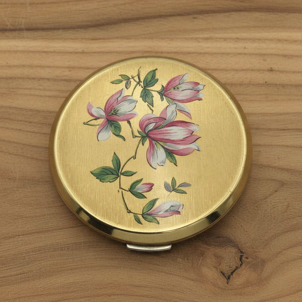 Brushed gold powder compact featuring pink and white flowers on the lid on wood